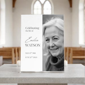 Könnte beinhalten: Gedenktafel mit dem Text „Celebrating the life of Emilia Watson“ und einem Schwarz-Weiß-Foto einer lächelnden Frau. Das Schild enthält die Geburts- und Sterbedaten der Verstorbenen. Das Schild wird auf einem Tisch angezeigt.