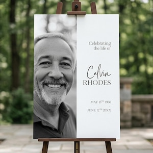 May include: A black and white memorial sign on a wooden easel. The sign features a portrait of a smiling man, the text "Celebrating the life of Calvin Rhodes," and dates: May 15th, 1960 - June 12th, 20XX.