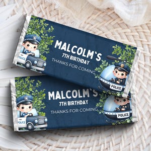 Op de afbeelding: Twee marineblauwe snoepreepverpakkingen met een politiethema. Elke verpakking toont een stripfiguur politieagent in een auto en helikopter, met de tekst "MALCOLM'S 7TH BIRTHDAY THANKS FOR COMING". Groene bladeren accentueren het ontwerp.
