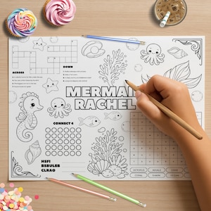 May include: A white paper placemat with a mermaid theme, featuring a crossword puzzle, Connect 4, and various sea creature illustrations. The text "MERMAI RACHEL" is prominently displayed. A hand is holding a pencil, ready to color. Two cupcakes and a drink are in the background.