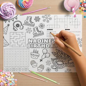 May include: A birthday-themed activity placemat with the text "Nadine's Birthday" is being colored with a pencil. The placemat features illustrations of ice skates, snowflakes, a cake, balloons, and a masquerade mask. Cupcakes and a drink are also visible.