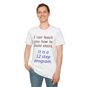 Può includere: Una t-shirt bianca a maniche corte con il testo in marrone scuro "I can teach you how to build stairs," seguito dal testo in blu "It is a 12 step program." La maglietta presenta un girocollo e una vestibilità standard in tessuto bianco.