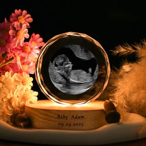 Op de afbeelding: Een kristallen bol met een echobeeld van een baby, rustend op een houten voet. De voet is gegraveerd met "Baby Adam" en de datum "09.09.2025". De bol is verlicht, met roze en gele bloemen en decoratieve veren op de achtergrond.