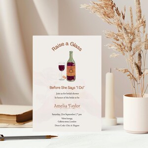 May include: Bridal shower invitation with the text "Raise a Glass" and "Before She Says 'I Do'". The invitation features a watercolour illustration of a wine bottle, a glass of wine, and a cork. The invitation also includes the name Amelia Taylor, date, time, location, and dress code.