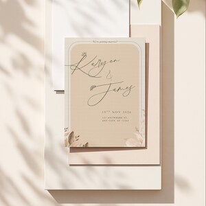 May include: A wedding invitation with a beige background and elegant script. The text reads "Karen & James" with the date "19th Nov 2026" and address details. Floral and crown illustrations add a decorative touch. The words "We're getting married!" are at the top.