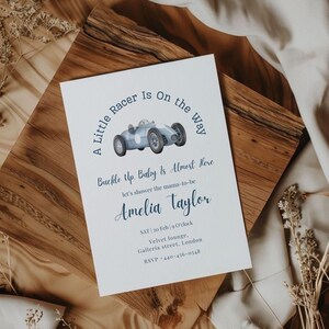 May include: A baby shower invitation featuring a blue vintage race car illustration. The text includes "A Little Racer Is On the Way" and "Buckle Up. Baby Is Almost Here." Details for Amelia Taylor's shower are included, with date, time, and location.