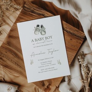 May include: A baby shower invitation with the text "A BABY BOY is almost here" and "Amelia Taylor". The invitation features watercolor illustrations of baby shoes and bunny rabbits. The invitation is on a white card, set on a wooden surface.