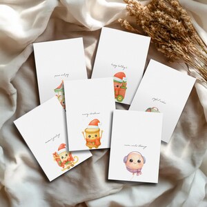 May include: A selection of white Christmas cards with watercolour illustrations and handwritten text. The cards feature festive designs such as a carousel, a gift, and a balloon character, with phrases like "peace and joy" and "happy holidays."