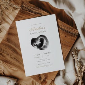May include: A baby shower invitation in white with gold lettering. The design features a watercolor image of a fetus within a heart. The invitation includes the text "Amelia's BABY SHOWER" and event details.