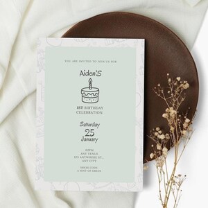 May include: A light green invitation card for Aiden's 1st birthday celebration. The card features a cake illustration and event details, including the date, time, and dress code. The card is on a brown plate with dried flowers.