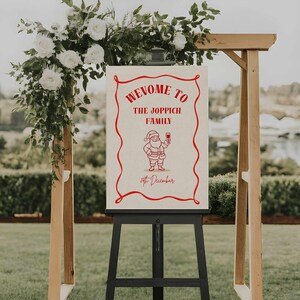 May include: A festive welcome sign on a black easel, featuring red text and a Santa illustration. The sign reads "Welcome to the Joppich Family" and the date "15th December". A wooden arch decorated with white flowers and foliage frames the sign, creating a welcoming display.