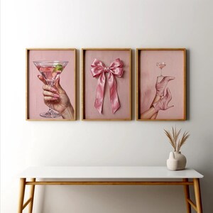 May include: Three framed art prints with a pink theme. One print features a hand holding a cocktail, another a pink bow, and the third a pair of pink boots holding a cocktail glass. The frames are wooden.