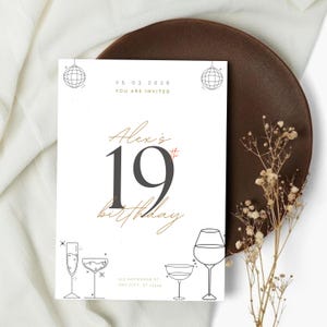 May include: A white birthday invitation card with gold and black text. The card reads "Alex's 19th birthday" with line drawings of glassware. The date "25.03.2026" is displayed at the top. The card is on a brown plate.