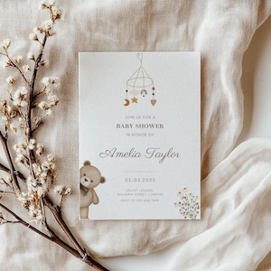 May include: A baby shower invitation in a cream and beige colour scheme. The design includes a teddy bear, a mobile with stars and moons, and floral details. The text reads "Join us for a Baby Shower in Honor of Amelia Taylor."