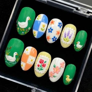 May include: A set of ten press-on nails in a clear plastic case. The nails feature various designs, including ducks, flowers, and checkered patterns. Colors include green, white, yellow, orange, and blue. The nails are oval-shaped.