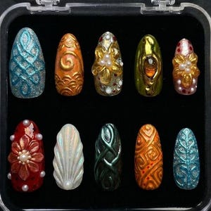 May include: A clear plastic case containing ten ornate, almond-shaped artificial nails. The nails feature various designs, including swirls, floral patterns, and embellishments in colors like gold, red, blue, and green. The nails are displayed against a black background.