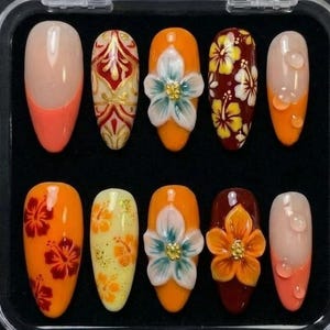 May include: A set of ten artificial nails in a clear plastic case. The nails feature various designs, including floral patterns, 3D flowers, and ombre effects in shades of orange, red, yellow, and white. Some nails have a French tip design.