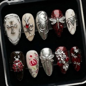 May include: A set of ten artificial nails in a clear case. The nails feature various designs, including crosses, dragonflies, floral patterns, and abstract silver embellishments. Colors include white, red, silver, and black, with a glossy finish.