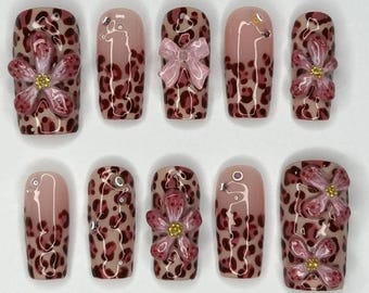 May include: A set of ten artificial nails with a leopard print design in shades of brown and tan. Some nails feature pink bows or 3D pink flowers with gold centers. The nails have a glossy finish.