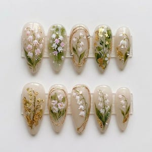 May include: A set of ten almond-shaped artificial nails. Each nail features a hand-painted design of lily of the valley flowers in white, pink, and green, with gold accents. Some nails have gold leaf details and small pearl embellishments.