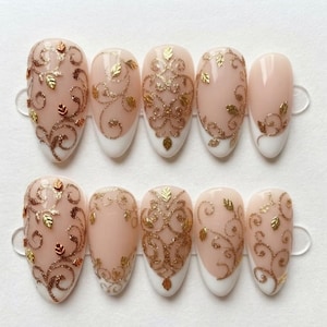May include: A set of ten almond-shaped press-on nails featuring a nude base with white tips. Each nail is decorated with intricate gold glitter swirls and small metallic gold leaf accents arranged in symmetrical and flowing patterns.