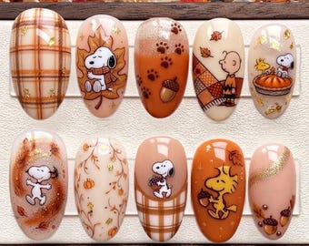 May include: A set of ten press-on nails featuring autumn-themed designs with Snoopy and Woodstock characters. The nails display patterns of plaid, falling leaves, pumpkins, acorns, and paw prints in shades of orange, brown, and cream with gold accents.