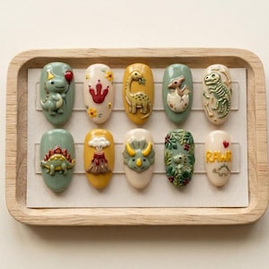 May include: A set of ten oval-shaped artificial nails with dinosaur-themed designs. Each nail features a different dinosaur or prehistoric element, including a baby dinosaur hatching from an egg, a volcano, and the word "RAWR". The nails are arranged on a wooden tray.