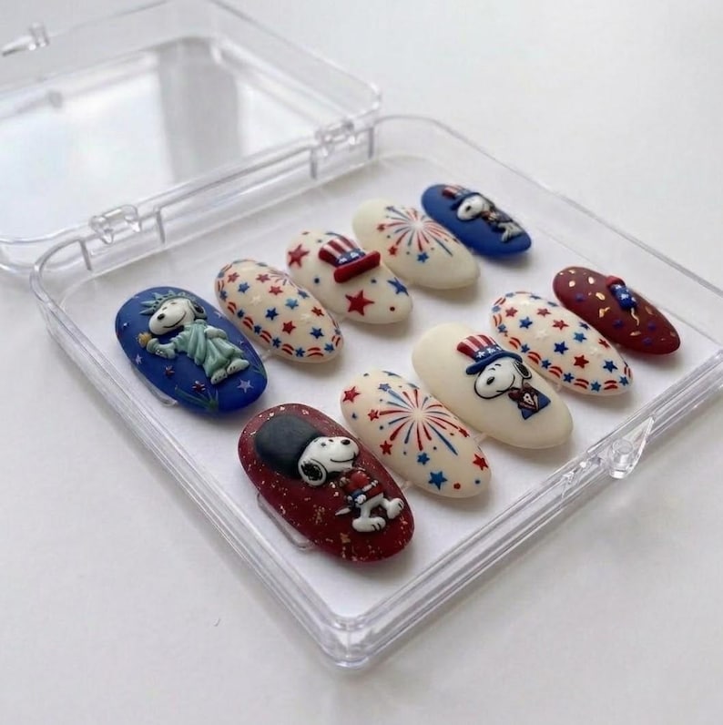 Pode incluir: Um conjunto de dez unhas posti&ccedil;as numa caixa de pl&aacute;stico transparente. As unhas apresentam desenhos patri&oacute;ticos com personagens Snoopy, estrelas e fogos de artif&iacute;cio em vermelho, branco e azul. Algumas unhas t&ecirc;m uma base vermelha escura ou azul, outras s&atilde;o branco sujo.