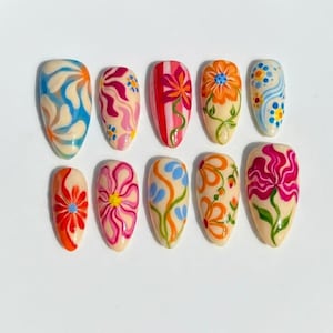 Puede incluir: Un conjunto de diez u&ntilde;as postizas en forma de almendra con base color crema y dise&ntilde;os pintados a mano, que incluyen remolinos azules y naranjas, patrones florales rosas y rojos, tallos verdes y puntos multicolores en dos filas.