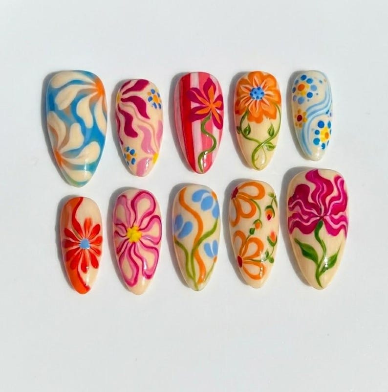 Puede incluir: Un conjunto de diez u&ntilde;as postizas en forma de almendra con base color crema y dise&ntilde;os pintados a mano, que incluyen remolinos azules y naranjas, patrones florales rosas y rojos, tallos verdes y puntos multicolores en dos filas.