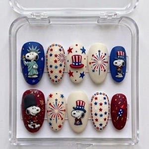 Pode incluir: Um conjunto de dez unhas artificiais numa caixa de pl&aacute;stico transparente. As unhas apresentam desenhos patri&oacute;ticos com as cores vermelho, branco e azul. Os desenhos incluem personagens Snoopy, estrelas, fogos de artif&iacute;cio e chap&eacute;us do Tio Sam.