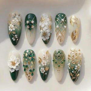 May include: A collection of ten almond-shaped artificial nails, each uniquely designed with a gradient of white to deep green. Embellishments include white flowers, gold accents, and green gemstones, creating an elegant, artistic aesthetic.