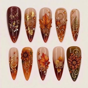 May include: A set of ten almond-shaped press-on nails featuring autumn-themed designs. The nails display deep burgundy, brown, and nude gradients adorned with 3D gold metallic swirls, intricate floral patterns, and detailed orange maple leaves.