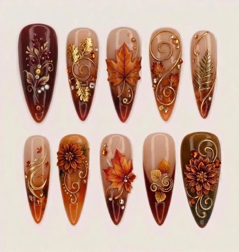 May include: A set of ten almond-shaped press-on nails featuring autumn-themed designs. The nails display deep burgundy, brown, and nude gradients adorned with 3D gold metallic swirls, intricate floral patterns, and detailed orange maple leaves.