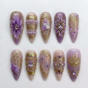 May include: A collection of ten ornate, long, almond-shaped artificial nails. Each nail features intricate designs, including 3D floral accents, gold filigree, and sparkling gems. The color palette includes shades of purple, pink, and gold.