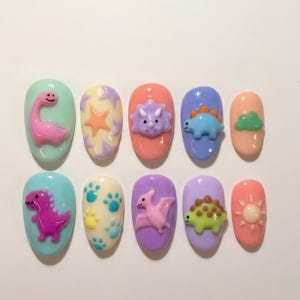 May include: A set of ten press-on nails featuring colorful 3D dinosaur and nature-themed charms. Designs include a pink long-neck dinosaur, purple stars, a triceratops head, a blue stegosaurus, a green cloud, a purple T-Rex, paw prints, a pterodactyl, a green ankylosaurus, and a sun.