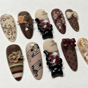 May include: A set of ten almond-shaped press-on nails featuring brown, cream, and black designs. Embellishments include 3D flowers, bows, musical notes, coffee beans, and swirling patterns. The collection showcases a mix of polka dots, stripes, and musical staff motifs in a cohesive color palette.