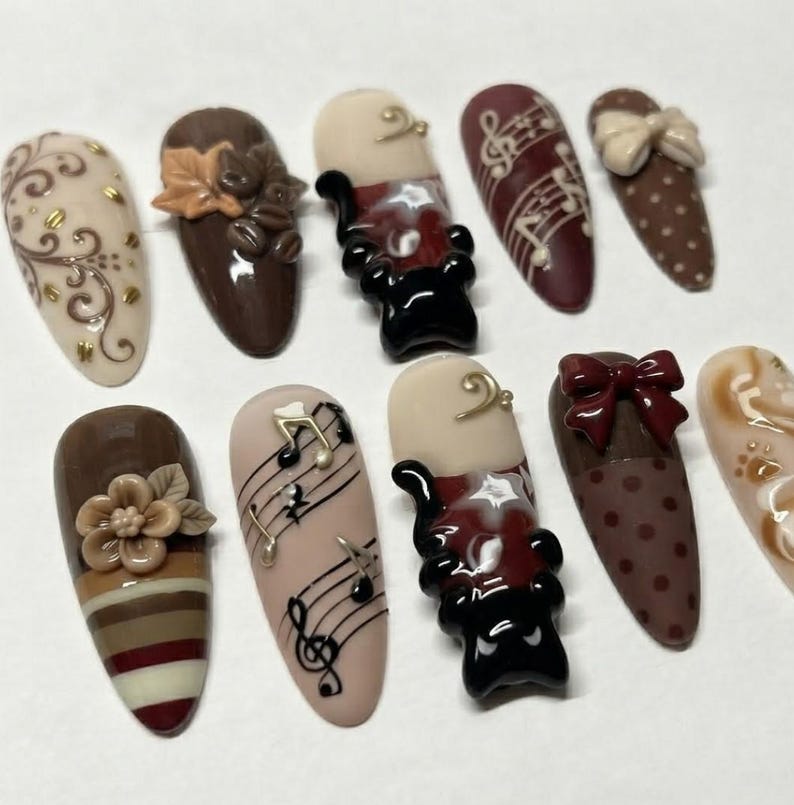 May include: A set of ten almond-shaped press-on nails featuring brown, cream, and black designs. Embellishments include 3D flowers, bows, musical notes, coffee beans, and swirling patterns. The collection showcases a mix of polka dots, stripes, and musical staff motifs in a cohesive color palette.