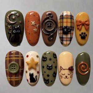 May include: A set of ten oval-shaped press-on nails featuring autumn-themed designs including black cats, crescent moons, stars, plaid patterns, buttons, paw prints, and bows in shades of olive green, orange, brown, cream, and yellow.
