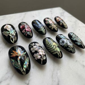 May include: A collection of ten black, oval-shaped artificial nails, each adorned with intricate floral designs. The nail art features various flowers in shades of pink, white, blue, and green, with iridescent accents. The nails are arranged on a white marble surface.