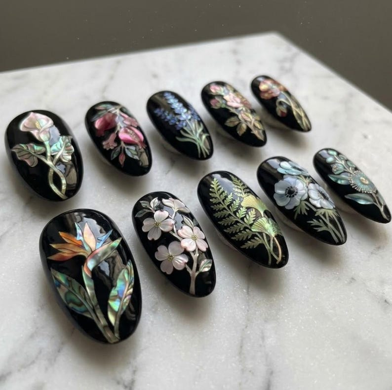 May include: A collection of ten black, oval-shaped artificial nails, each adorned with intricate floral designs. The nail art features various flowers in shades of pink, white, blue, and green, with iridescent accents. The nails are arranged on a white marble surface.