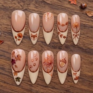 May include: A set of ten almond-shaped press-on nails featuring a nude base with copper-colored French tips. The designs include autumn-themed illustrations such as a fox face, paw prints, an acorn, various fall leaves, and floral patterns.