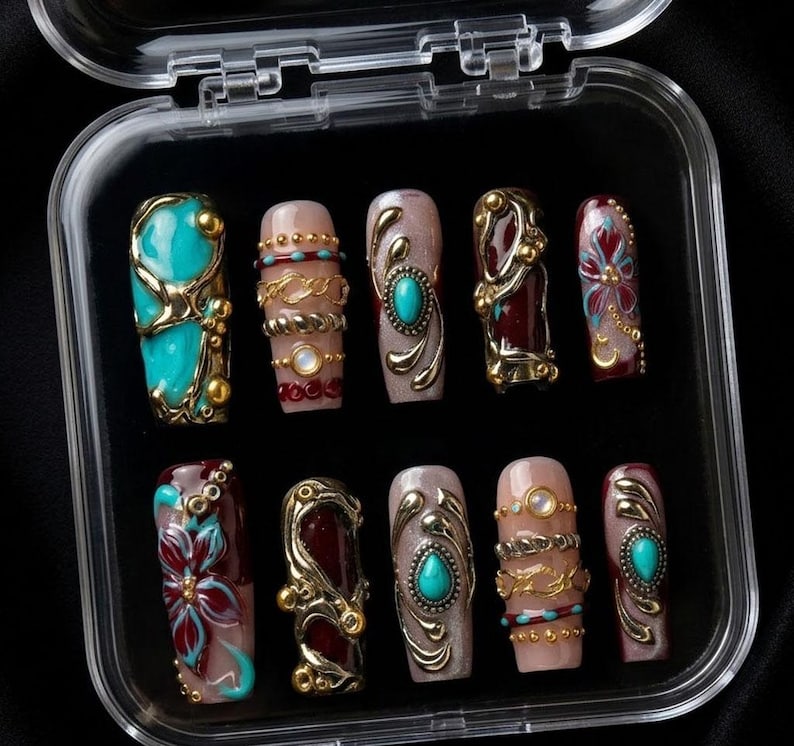 May include: A set of ten artificial nails in a clear plastic case. The nails feature various designs, including turquoise stones, gold embellishments, and floral patterns. The nails are in shades of teal, burgundy, and nude.