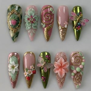 May include: A collection of ten long, pointed artificial nails with intricate designs. The nails feature various colors, including pink, green, and gold, and are adorned with 3D floral, fruit, and insect embellishments. Some nails have gold accents and glitter.