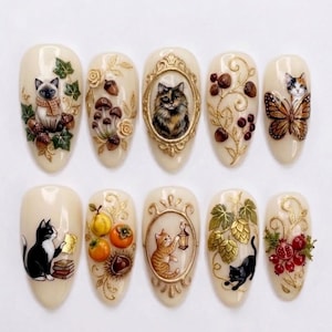 May include: A set of ten almond-shaped press-on nails with a cream base, featuring intricate 3D-style designs including cats, gold-toned ornate frames, mushrooms, acorns, butterflies, autumn fruits, lanterns, and botanical elements like leaves and berries.