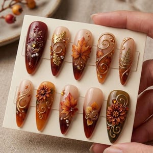 May include: A set of ten almond-shaped press-on nails featuring autumn-themed designs. The nails display warm colors like deep red, orange, and brown, decorated with 3D gold leaf accents, floral motifs, swirling lines, and small embedded crystals.