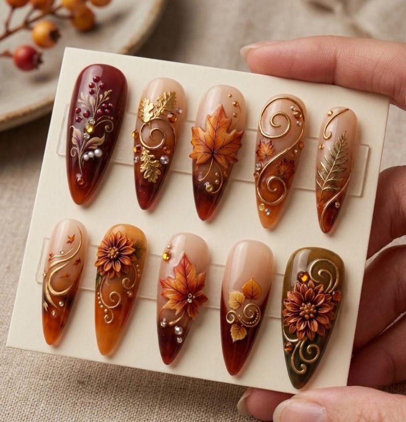 May include: A set of ten almond-shaped press-on nails featuring autumn-themed designs. The nails display warm colors like deep red, orange, and brown, decorated with 3D gold leaf accents, floral motifs, swirling lines, and small embedded crystals.