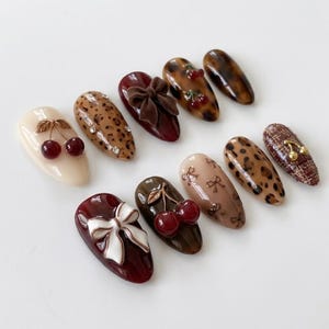 K&ouml;nnte beinhalten: Ein Set aus zehn mandelf&ouml;rmigen Press-on-N&auml;geln mit verschiedenen Designs, darunter cremefarbene, burgunderrote und leopardenmusterte Basen. Verzierungen umfassen 3D-rote Kirsch-Charms, wei&szlig;e und braune Schleifen, kleine Strasssteine und goldene Kirschen.