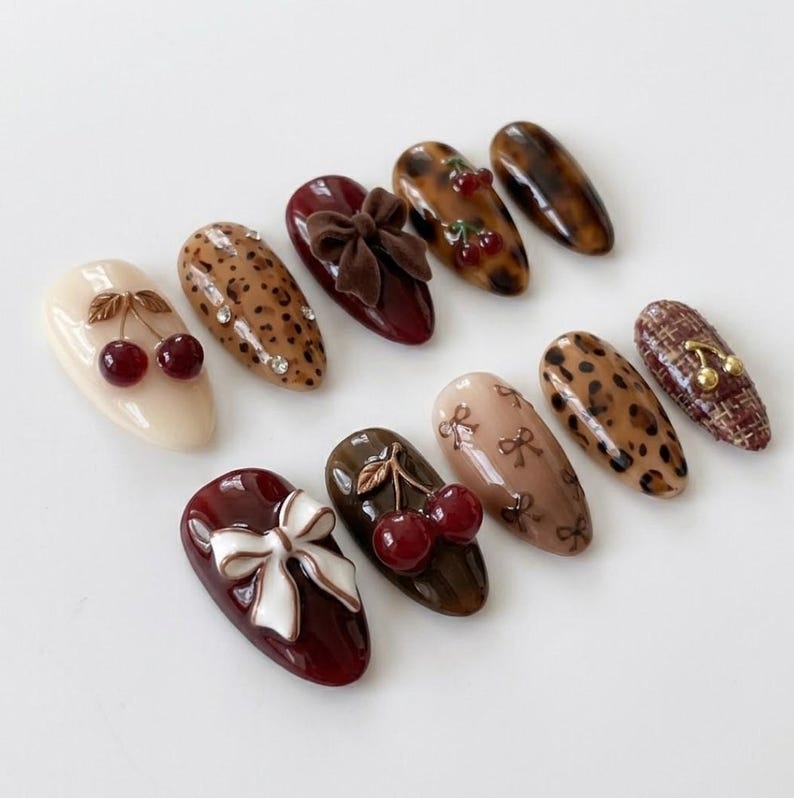 K&ouml;nnte beinhalten: Ein Set aus zehn mandelf&ouml;rmigen Press-on-N&auml;geln mit verschiedenen Designs, darunter cremefarbene, burgunderrote und leopardenmusterte Basen. Verzierungen umfassen 3D-rote Kirsch-Charms, wei&szlig;e und braune Schleifen, kleine Strasssteine und goldene Kirschen.