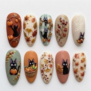 May include: A set of ten almond-shaped press-on nails featuring autumn-themed designs. The collection includes illustrations of black cats, orange pumpkins, brown and yellow fall leaves, woven baskets, and delicate white lace patterns on various backgrounds.